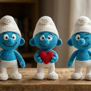 May include: Three handmade crochet Smurf figurines. Each Smurf is blue with white pants and a white hat. One Smurf holds a red heart. The figurines are displayed on a wooden surface.