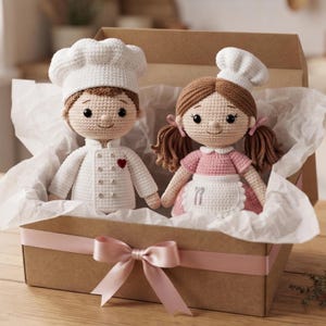 May include: Two crocheted dolls inside a cardboard gift box with a pink ribbon. One doll wears a white chef hat and jacket with a red heart. The other doll has brown pigtails, a white chef hat, a pink dress, and a white apron with a fork and spoon icon.
