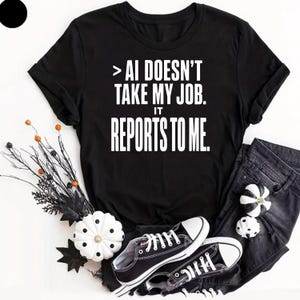Könnte beinhalten: Ein schwarzes Kurzarm-T-Shirt mit dem weißen Aufdruck > AI DOESN'T TAKE MY JOB. IT REPORTS TO ME. Das T-Shirt wird zusammen mit schwarzem Jeansstoff, schwarz-weißen Turnschuhen sowie dekorativen schwarz-weißen Kürbissen präsentiert.