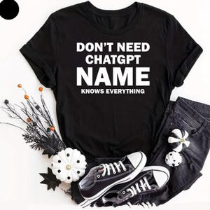 May include: A black short-sleeved t-shirt featuring the white text DON'T NEED CHATGPT NAME KNOWS EVERYTHING. The shirt is displayed alongside black denim trousers, black and white trainers, and decorative white pumpkins with black polka dots.