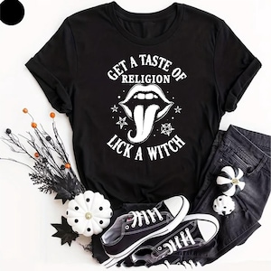 May include: Black t-shirt with white text that reads "GET A TASTE OF RELIGION LICK A WITCH" and a graphic of a mouth with a long tongue. The shirt is paired with black jeans and black and white sneakers. Decorative pumpkins and branches are also in the image.