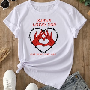 May include: A white short-sleeve t-shirt featuring a graphic print of two red hands forming a heart shape inside a black barbed wire heart. The text "SATAN LOVES YOU" appears above the graphic, and "FOR WHO YOU ARE" is printed below in red.