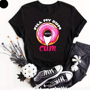 May include: A black short-sleeved t-shirt featuring a graphic of a pink frosted donut with colourful sprinkles. The text "FILL MY BUM" is printed in white above the donut, "WITH" is in the centre, and "cum" is in pink below the donut.