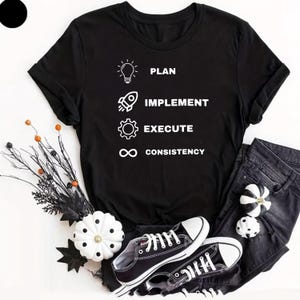 May include: A black short-sleeved t-shirt featuring a white graphic print with four icons and corresponding text: a lightbulb labelled PLAN, a rocket labelled IMPLEMENT, a gear labelled EXECUTE, and an infinity symbol labelled CONSISTENCY.