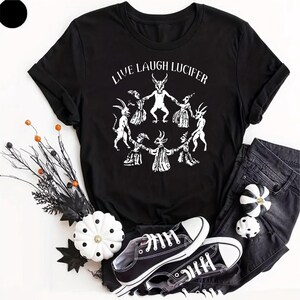 May include: A black short-sleeved t-shirt featuring a white graphic print of figures in a circle with the text "LIVE LAUGH LUCIFER" above them. The shirt is displayed alongside black denim trousers, black and white trainers, and pumpkins.