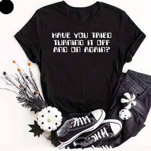 May include: A black short-sleeved t-shirt with the white text "HAVE YOU TRIED TURNING IT OFF AND ON AGAIN?" printed on the front. The shirt is displayed alongside black denim jeans, black and white sneakers, and decorative pumpkins.