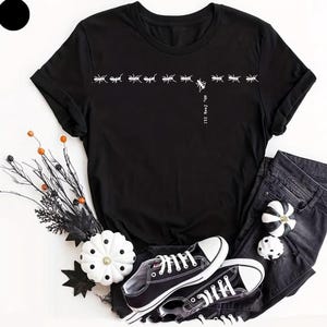 May include: A black short-sleeved t-shirt featuring a horizontal line of white ant illustrations across the chest. One ant is separated from the line, accompanied by the white text "ah, fook it!" printed vertically below it in a simple font.