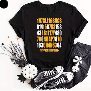 Puede incluir: Una camiseta negra de manga corta con un estampado gráfico de texto alfanumérico amarillo y blanco que dice 1N73LL163NC3, 0141587H3158, 43481L17Y480, 7004D4P7870, 183CH4N6304 y -573PH3N H4WKING- en un diseño de bloque vertical.