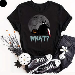 May include: A black short-sleeved t-shirt featuring a graphic of a black cat holding a bloody knife next to a jack-o'-lantern in front of a full moon with bats. The text "WHAT?" is printed below in a light blue, dripping, horror-style font.