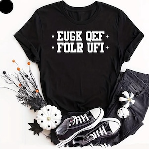 May include: A black short-sleeved t-shirt featuring the white text EUGK QEF FOLR UFI printed on the chest. The shirt is displayed alongside black denim jeans, black and white trainers, and decorative white pumpkins with black polka dots.