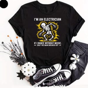 May include: A black t-shirt featuring a white skeleton graphic surrounded by yellow lightning bolts. The text reads: I'M AN ELECTRICIAN IF I DANCE WITHOUT MUSIC. SHUT THE MAIN BREAKER OFF. The shirt is displayed with black jeans and black sneakers.