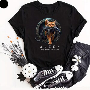 May include: A black short-sleeved t-shirt featuring a graphic of an orange tabby cat carrying a black xenomorph creature in its mouth, set within a circular frame. Below the graphic, the text reads ALIEN THE SHORT VERSION in white capital letters.