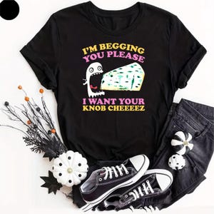 May include: A black short-sleeved t-shirt featuring a graphic of a white ghost with a wide-open mouth eating a wedge of blue cheese. The text reads I'M BEGGING YOU PLEASE I WANT YOUR KNOB CHEEEEZ in yellow and pink lettering on the front.