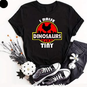 Puede incluir: Camiseta negra con el texto "I Raise Dinosaurs Tiny" en un diseño de logotipo de película. El gráfico presenta un círculo rojo con una silueta de gallina y polluelos. La camiseta se combina con vaqueros negros y zapatillas deportivas blancas y negras.