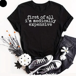 May include: A black short-sleeved t-shirt featuring the white text "first of all i'm medically expensive" in a typewriter-style font. The shirt is paired with black denim jeans and black and white canvas trainers, accented by decorative pumpkins.
