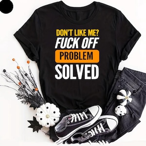 Könnte beinhalten: Schwarzes T-Shirt mit dem Text "DON'T LIKE ME? FUCK OFF PROBLEM SOLVED" in Gelb, Orange und Weiß. Das Shirt wird mit einer schwarzen Jeans und schwarz-weißen Turnschuhen kombiniert. Dekorative Kürbisse und Zweige sind ebenfalls im Bild.