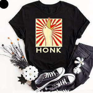 May include: Black t-shirt featuring a cartoon goose with an open beak, set against a red and cream background with the word "HONK" below. The shirt is paired with black jeans and black and white sneakers.