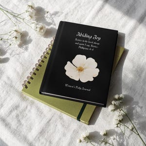 May include: A black journal with the text "Abiding Joy" and a white flower design. The journal also includes the text "Rejoice in the Lord alway: and again I say, Rejoice. Phillipians 4:4" and "Women's Daily Journal". A green spiral notebook is beneath it.