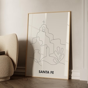 May include: A framed wall art print featuring a black continuous line drawing of a mission church with a cross, a vase, and a plant on a light beige background. The text SANTA FE is printed in bold black capital letters at the bottom of the print.