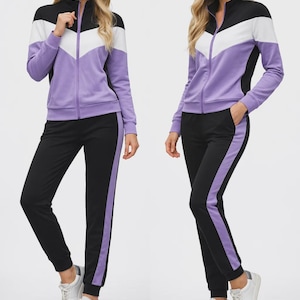 Könnte beinhalten: Ein zweiteiliger Trainingsanzug in Schwarz, Weiß und Lila. Die Jacke hat einen schwarzen Kragen und Ärmel, ein weißes Chevron-Design und einen lilafarbenen Körper. Die Hose ist schwarz mit einem lila Streifen an der Seite. Das Model trägt weiße Turnschuhe.