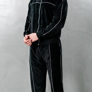 K&ouml;nnte beinhalten: Schwarzer Velours-Trainingsanzug mit wei&szlig;em Paspel. Die Jacke hat einen durchgehenden Rei&szlig;verschluss und die Hose hat ein gerades Bein. Das Model tr&auml;gt wei&szlig;e Turnschuhe. Dieser Trainingsanzug ist ein bequemes und stilvolles Sportoutfit.