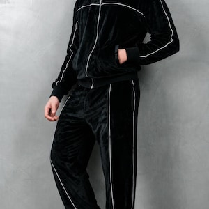 K&ouml;nnte beinhalten: Schwarzer Velours-Trainingsanzug mit wei&szlig;er Paspelierung. Die Jacke hat einen Rei&szlig;verschluss und Taschen, und die Hose hat einen geraden Beinverlauf. Das Outfit wird mit wei&szlig;en Turnschuhen getragen. Dieser bequeme Trainingsanzug ist f&uuml;r Freizeitkleidung geeignet.