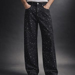 Puede incluir: Pantalones vaqueros negros de pernera ancha, adornados con numerosos brillantes en forma de estrella. Los pantalones tienen un dise&ntilde;o cl&aacute;sico de cinco bolsillos y cierre de bot&oacute;n y cremallera. El modelo lleva zapatos negros y una camiseta sin mangas blanca.