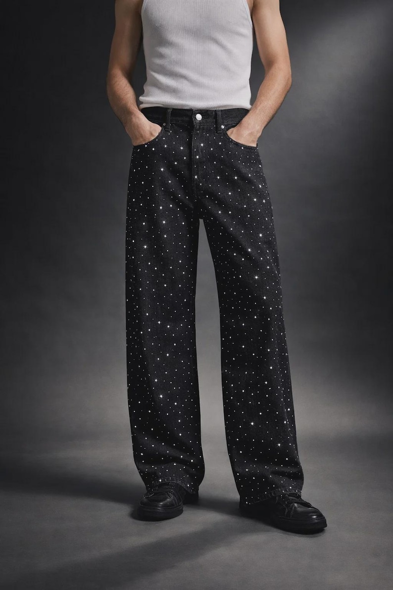 Puede incluir: Pantalones vaqueros negros de pernera ancha, adornados con numerosos brillantes en forma de estrella. Los pantalones tienen un dise&ntilde;o cl&aacute;sico de cinco bolsillos y cierre de bot&oacute;n y cremallera. El modelo lleva zapatos negros y una camiseta sin mangas blanca.