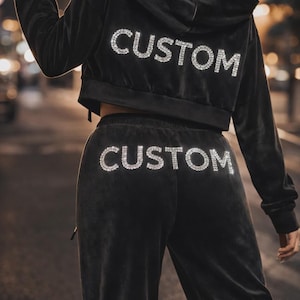 Könnte beinhalten: Schwarzes Velours-Hoodie und passendes Hosen-Set. Das Wort "CUSTOM" ist mit glitzernden Strasssteinen auf der Rückseite des Hoodies und auf der Hose geschrieben. Das Outfit ist ein Cropped-Style.