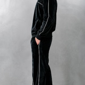 K&ouml;nnte beinhalten: Ein schwarzer Velours-Trainingsanzug mit wei&szlig;en Paspeln. Der Anzug besteht aus einer Jacke mit Rei&szlig;verschluss und einer passenden Hose. Die Person tr&auml;gt wei&szlig;e Turnschuhe. Das Outfit ist ein l&auml;ssiger, bequemer Stil.