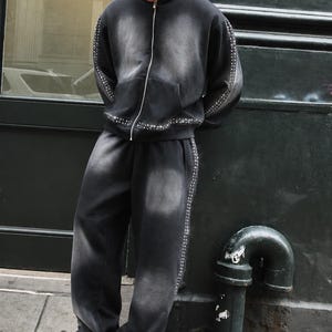 Op de afbeelding: Een zwarte hoodie en joggingbroek set met denim-wash en zilveren stud accenten. De hoodie heeft een ritssluiting aan de voorkant en een capuchon met trekkoord. De joggingbroek heeft een relaxte pasvorm. Het model draagt witte sneakers.