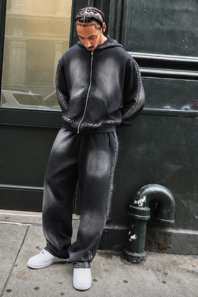 Op de afbeelding: Een zwarte hoodie en joggingbroek set met denim-wash en zilveren stud accenten. De hoodie heeft een ritssluiting aan de voorkant en een capuchon met trekkoord. De joggingbroek heeft een relaxte pasvorm. Het model draagt witte sneakers.