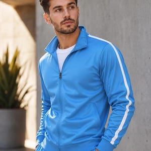 Puede incluir: Un chándal azul claro con rayas blancas en las mangas y las piernas. La chaqueta tiene cremallera y cuello alzado. Se lleva una camiseta blanca debajo. El chándal está hecho de un material liso y brillante.