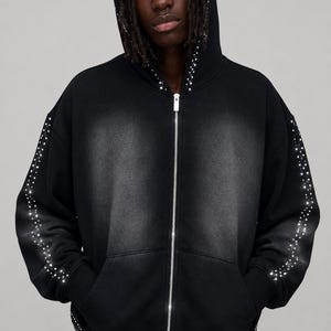 Op de afbeelding: Zwarte hoodie met ritssluiting en een ontwerp met studs. De hoodie heeft een zilveren rits en is versierd met kleine, glinsterende studs langs de capuchon, mouwen en voorkant. Het kledingstuk heeft een vervaagde, versleten uitstraling.