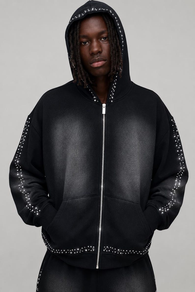 Op de afbeelding: Zwarte hoodie met ritssluiting en een ontwerp met studs. De hoodie heeft een zilveren rits en is versierd met kleine, glinsterende studs langs de capuchon, mouwen en voorkant. Het kledingstuk heeft een vervaagde, versleten uitstraling.