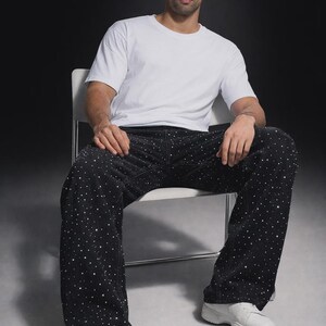 Puede incluir: Una persona sentada en una silla con una camiseta blanca, pantalones negros con adornos de puntos blancos y zapatillas blancas. El fondo es gris oscuro.