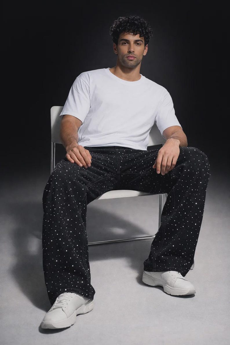 Puede incluir: Una persona sentada en una silla con una camiseta blanca, pantalones negros con adornos de puntos blancos y zapatillas blancas. El fondo es gris oscuro.