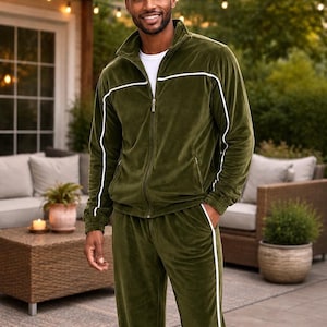 May include: Olive green velour track suit with white trim. The jacket has a zip-up front and a stand-up collar. The pants have a straight leg design. The outfit is worn by a person standing outside.