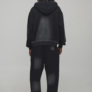 Op de afbeelding: Zwarte hoodie en bijpassende broek. De hoodie heeft een geplooide rand langs de capuchon en zoom. De broek heeft ook geplooide accenten bij de enkels. De outfit is een donkere wassing denim stijl.