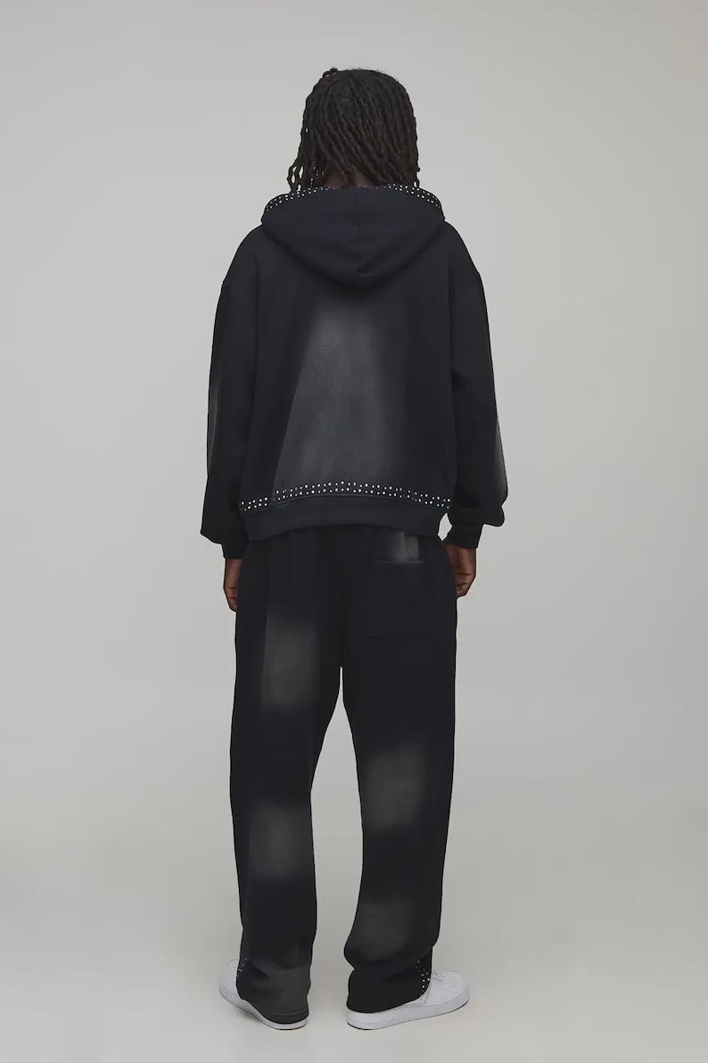 Op de afbeelding: Zwarte hoodie en bijpassende broek. De hoodie heeft een geplooide rand langs de capuchon en zoom. De broek heeft ook geplooide accenten bij de enkels. De outfit is een donkere wassing denim stijl.