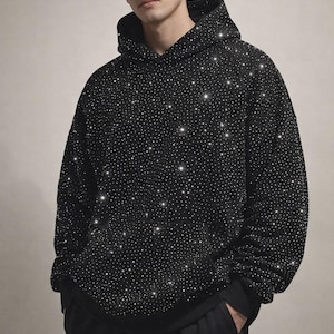 May include: Black hoodie covered in small, sparkling embellishments. The hoodie has a drawstring hood, long sleeves, and a kangaroo pocket. The person is wearing black pants.