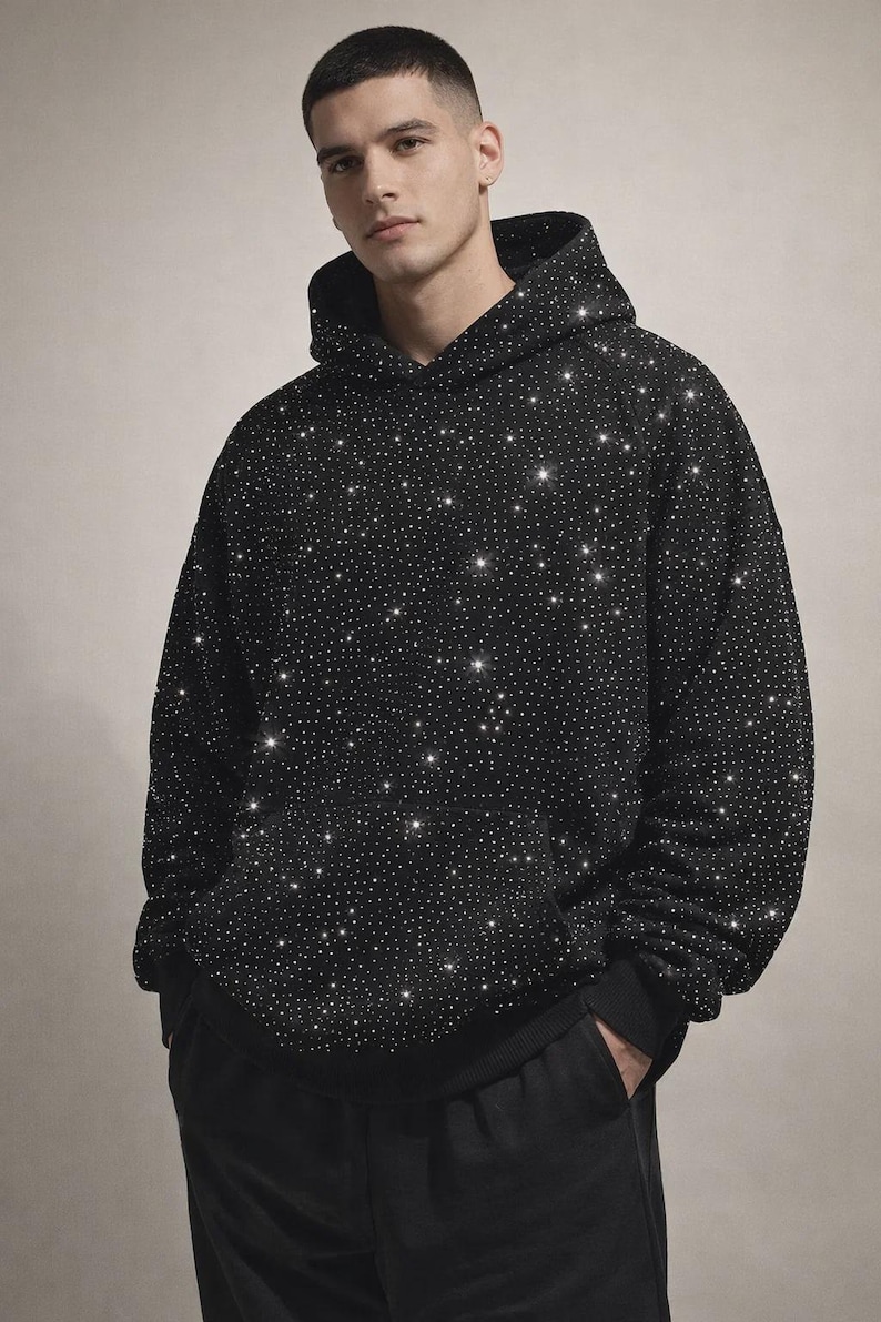 May include: Black hoodie covered in small, sparkling embellishments. The hoodie has a drawstring hood, long sleeves, and a kangaroo pocket. The person is wearing black pants.