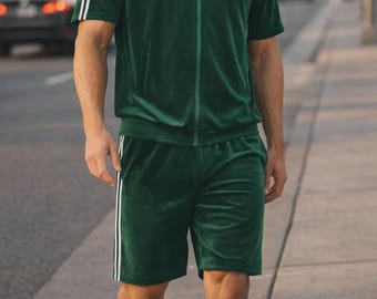 May include: A green velvet tracksuit, featuring a zip-up jacket with white stripes on the sleeves and matching shorts. The shorts also have white stripes down the sides. The outfit is paired with green sneakers.