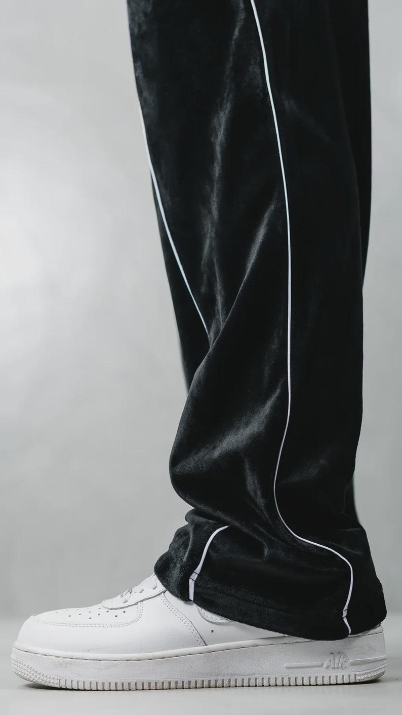 K&ouml;nnte beinhalten: Schwarze Samt-Trainingshose mit wei&szlig;em Paspeldetail. Die Hose wird mit wei&szlig;en Ledersneakern kombiniert. Die Sneaker haben das Wort "AIR" auf der Seite. Die Hose ist leicht zerknittert.