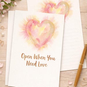 May include: Two white cards featuring watercolour heart designs in pink, yellow, and peach hues. The text "Open When You Need Love" is printed on one card. A wooden pen and decorative elements are also present.