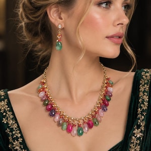 Puede incluir: Un conjunto de collar y pendientes colgantes en tono dorado con piedras preciosas facetadas en forma de lágrima en tonos rosa, verde y morado, acentuadas con pequeñas cuentas rojas y cristales transparentes en una delicada cadena dorada.
