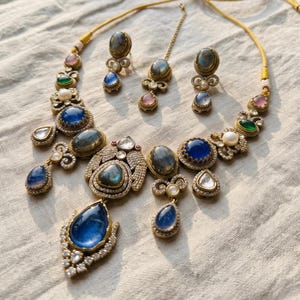 May include: A jewelry set featuring a necklace, two earrings, and a maang tikka. The pieces are crafted with gold-toned metal, set with blue, green, and pink cabochon stones, iridescent labradorite, white pearls, and clear crystals.