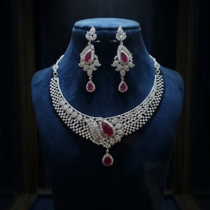 May include: A jewelry set consisting of a necklace and matching drop earrings. The necklace features a wide, silver-toned band encrusted with clear stones, a central teardrop-shaped red stone, and a small dangling red teardrop stone. The earrings mirror this design.