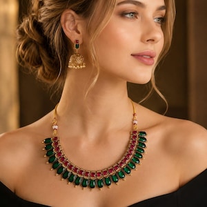 Puede incluir: Un collar de tono dorado con una hilera de piedras verdes en forma de lágrima acentuadas por una hilera de piedras rosas cuadradas, junto con pendientes jhumka a juego en forma de campana con una piedra verde y base de filigrana dorada.