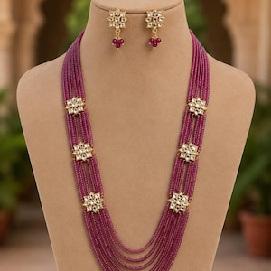 May include: A multi-strand necklace featuring deep magenta beads arranged in six layers, accented by three gold-toned floral motifs on each side set with clear stones. Matching stud earrings feature a single floral motif with three dangling magenta beads.